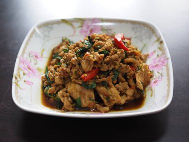 Tayland usulü kızarmış et, acı biber ve fesleğen. Çiçekli bir porselen tabakta servis ediliyor. Canlı renkler ve zengin dokular sergileniyor..