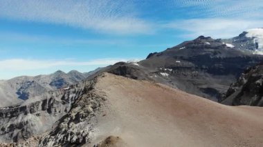 Yavaş bir yatay süzülüş And arazisinin ham büyüklüğünü ortaya çıkarır. Pürüzlü tepeler, kumlu tepeler ve uzak buzul tepeleri ince bulutlarla kaplanmış bir gökyüzünün altındaki çerçeveye uzanıyor..