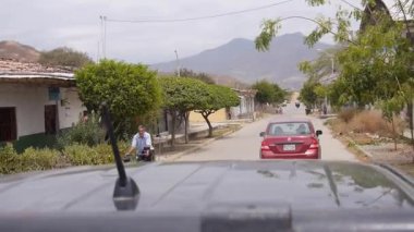 Bu video, Kuzey Peru 'daki tipik bir yerleşim yerini gösteriyor. Kırmızı bir sedan' ı takip eden bir kamyonetin yatağında görülüyor. Manzarada ağaçlar, mütevazı evler, elektrik hatları ve bulutlu bir gökyüzünün altındaki dağlar yer alıyor..