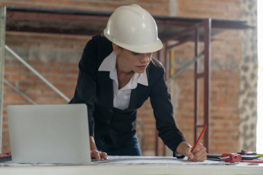 Profesyonel kadın mimar inşaat alanının planlarını inceliyor, başlık takıyor ve dizüstü bilgisayar kullanıyor..