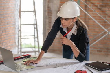 Güvenlik kaskı takmış odaklanmış bir kadın mimar bir inşaat alanının planlarını inceler, referans olarak bir dizüstü bilgisayar kullanır..