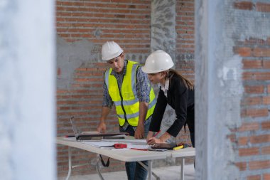 Güvenlik teçhizatı incelemesi yapan iki inşaat işçisi, tuğla duvarları ve beton sütunları olan bir inşaat sahasının planlarını inceliyor..