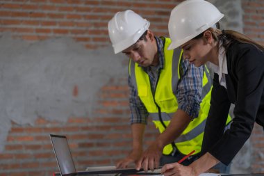 Güvenlik teçhizatı ve sert şapkalı iki inşaat işçisi endüstriyel bir ortamda dizüstü bilgisayarın planlarını gözden geçiriyorlar..