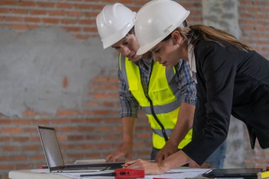 Bina planlarını inceleyen ve inşaat alanında dizüstü bilgisayar kullanan iki uzman..