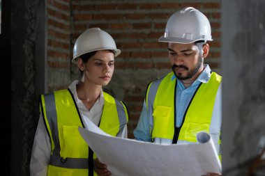 Güvenlik teçhizatı ve yüksek görüşlü yelekli iki inşaat işçisi bir inşaat alanının planlarını inceliyor..