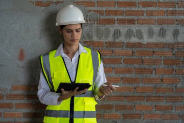 Yüksek görüş açısına sahip yelek ve başlık takan kadın inşaat işçisi. İnşaat alanında tuğladan bir arka planı olan tablet kullanıyor..