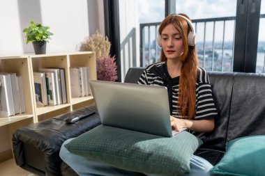 Uzun kızıl saçlı genç bir kadın kanepede oturuyor, dizüstü bilgisayarla çalışıyor. Kulaklık takıyor ve işine odaklanıyor. Etrafı bitkiler ve kitaplarla çevrili modern yaşam alanlarıyla çevrili.