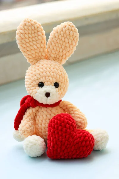 Saint Valentine\'s day creative image with a handmade teddy rabbit sitting with a big knitted red heart as a present. Self made knitted rabbit toy in pastel colors holding a red heart. Love and couple relationships. Cute plush rabbit toy as a present