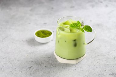 Iced matcha latte on a coaster with a small bowl of matcha powder on the back, gray background. Refreshing antioxidant natural drink made of ground powder of green tea leaves with milk, ice and fresh mint. Matcha green tea