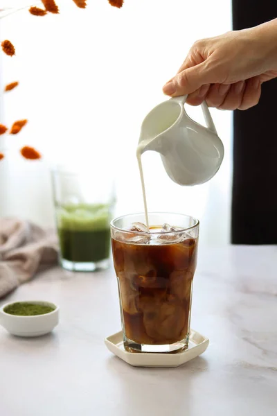 A transparent glass of iced black coffee with a hand holding a milk jug and pouring milk into coffee glass. Sunny morning with cold coffee and green matcha tea on a dining table, natural lights, light blurred background. Cozy home interior atmosphere