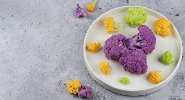 Yellow, purple and green fresh cauliflower inflorescences on white plate on grey background. Cutted raw cauliflower prepared to be cooked or ready to eat as raw food diet. Copy space for text. Rainbow in a plate