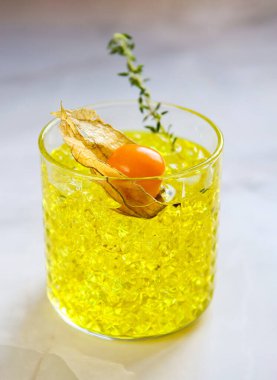 Yellow cocktail with physalis and a branch of thyme in a transparent glass on a light blurred background in natural daylights. Physalis cocktail with crushed ice on a marble table. Close up of a yellow drink. Cocktail with yellow liquor, soda and ice