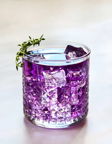 Purple drink made of violet liquor in a transparent drinking glass with ice cubes and a thyme sprig on the top. Violet cocktail. Iced colorful drink close up, isolated on a light grey blurred background in natural counterlight