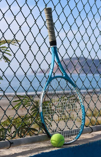 Tennis racket and tennis ball on the sandy beach in front of the sea and mountain on a sunny day. Summer sport concept. Tennis training by the sea. Tennis on the beach background. Workout at the seashore