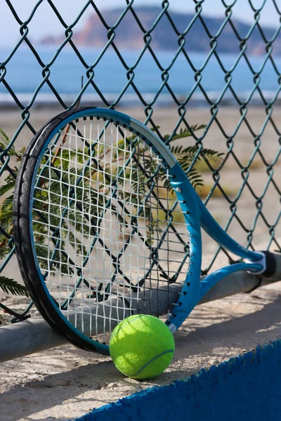 Tennis racket and tennis ball on the sandy beach in front of the sea and mountain on a sunny day. Summer sport concept. Tennis training by the sea. Tennis on the beach background. Workout at the sea shore