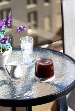 Güneşli bir öğleden sonra, üst kattaki bir kafede şeffaf bir masanın üzerinde bir bardak buzla bir fincan kahve ya da sıcak çikolata, bulanık şehir manzaralı. Çikolatalı kahve, akrabalar.