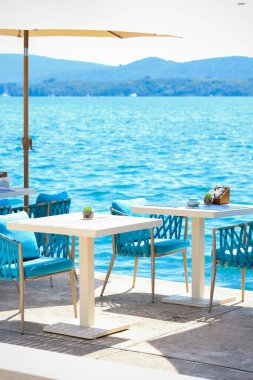 Plaj kulübü konsepti. Sahilde şık bir yemek köşesi ya da sahil kafesi. Restoran masaları, saman doğal sandalyeler, beyaz ahşap masalar, örgü sandalyeler, arka planda deniz manzarası.