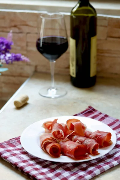 Plate with sliced prosciutto and a glass of red wine on kitchen table, lifestyle photography. Prsut and red wine, italian or spanish cured meats. Jamon appetizer with a glass of wine as brunch