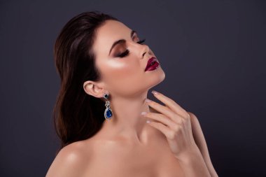 Gri arka planda izole edilmiş, boynundaki değerli küpelere dokunan göz kamaştırıcı, dumanlı gözlerin yan fotoğrafı..