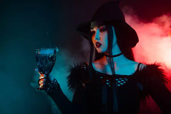 令人惊艳的邪恶女巫用高脚杯在雾蒙蒙的霓虹灯背景下准备神奇的黑色液体药水 — 图库照片