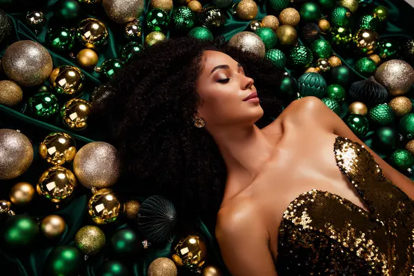 Portrait of stunning party lady lying on emerald silky texture covered decorated with xmas tree glitter baubles.