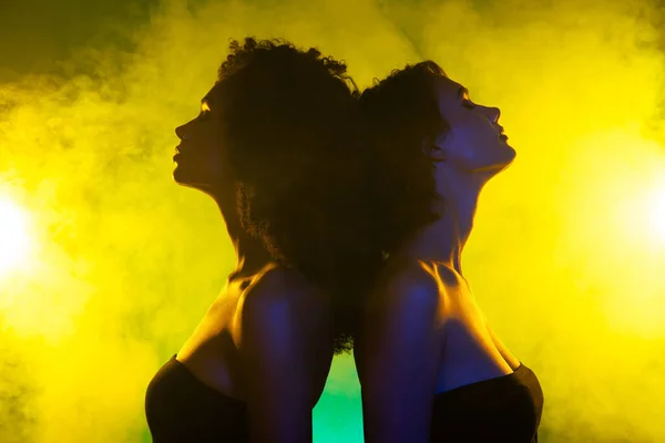 Photo of classy chic girls stand back to back couple dancing in night club over bright shine mist neon background.