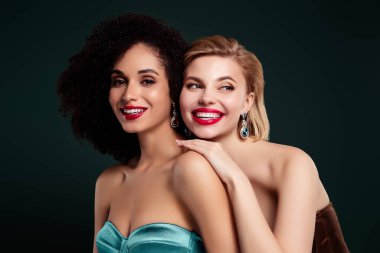 Photo of joyful pretty girls smiling touch shoulders look over dark green color empty space background.
