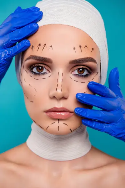 女性のプラスチック手術患者の肖像画は シアンバックグラウンド上の青い医療手袋で化粧品学者のタッチフェイスを見ます — ストック写真