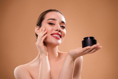 Neşeli sevimli kızın fotoğrafı merhem kavanozu tut, yüzüne dokun, bej rengi arka planda izole krem sür..