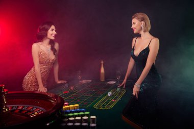 Yeşil masada poker oynayan iki kadının fotoğrafı. Yığınla fiş ve neon arka planda birbirlerine bakıyorlar..