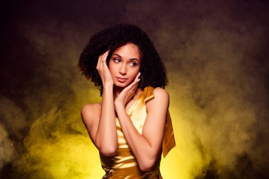 Gülümseyen, hayalperest, altın elbiseli bir kadının fotoğrafı. İzole edilmiş sarı ışık arka planında eğleniyor..