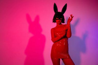Çekici bir kadının fotoğrafı eldiven giyip neon ışıklı arka planda izole edilmiş tavşan maskesi kırmızı deri giysiler giyiyor..