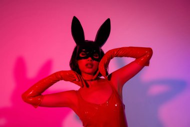 Göz kamaştırıcı bir kızın fotoğrafı yumuşak tenli tavşan maskeli kırmızı deri elbise neon ışıklı arka planda izole edilmiş..