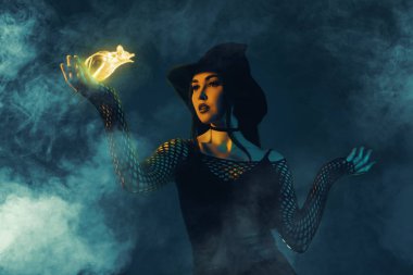 Cadı kostümlü kadının yaratıcı koyu renk kolaj posteri. El ele tutuşup sisli arkaplanı sarı yarasa büyüsüyle kaplamış..