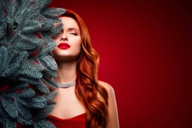 Çekici bir bayanın fotoğrafı, kırmızı arka planda gözleri kapalı bir şekilde yılbaşı ağacı dalının yanında saklanmayı cezbedici bulur..