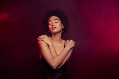 Çekici, güzel mankenin koyu kırmızı arka planda kendini kucaklayan yumuşak dokunuşunun fotoğrafı..