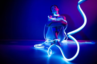 Koyu mavi arka planda parlayan neon ışıkların arasında oturan genç bir kadın, sanatsal bir stüdyoda teknolojik bir çarpıklıkla güzellik ve modern stil yayıyor..