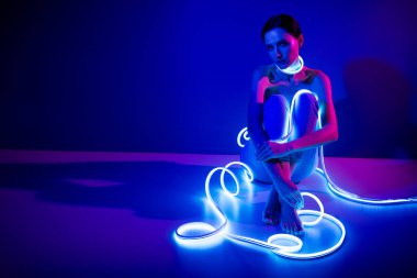 Genç bir kadın parıldayan neon şeritli bir sanat stüdyosunda poz veriyor. Koyu mavi arka plan onun güzelliğini ve tarzını vurguluyor, çağdaş ve çekici bir atmosfer yaratıyor..