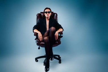 Siyah ceketli şık bir genç kadın, mavi bir arka planda deri bir sandalyede güven içinde oturuyor. Duruşu zarif ve güçlü..