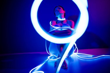 Genç bir kadın, parlak neon bir şeritle koyu mavi bir stüdyoda poz veriyor, fütüristik tarzı ve zarafeti kucaklıyor. Kendine güveni güzelliği ve modern modayı yansıtıyor..