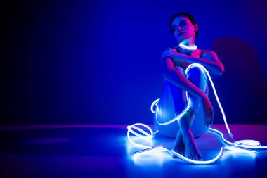 Genç bir kadın parlak neon şeritlerle çevrili, koyu mavi bir arkaplanda, fütüristik ve şık bir atmosfer yaratıyor..