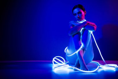 Neon ışıklarla kaplanmış genç bir kadın koyu mavi bir arka planda oturuyor, sanatsal bir stüdyo ortamında ihtişam ve zarafet yayıyor..