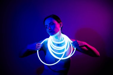 Genç bir kadın omuzlarının etrafında parlak neon bir şeritle koyu mavi arka plana karşı zarif poz veriyor. Görüntü güzellik, moda ve teknoloji içeriyor..