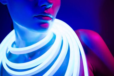 Omuzlarında parlak neon şeritlerle zarif genç bir kadın, koyu mavi bir arka plana karşı güzellik ve tarz yayıyor, çağdaş ve çekici bir moda özünü yakalıyor..