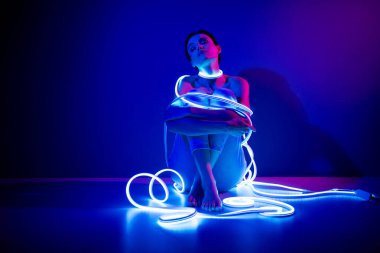 Büyüleyici bir kadın, neon ışıklara sarılmış, koyu mavi arka planda otururken, stüdyo ortamında zarafet ve modern stil sergiliyor..