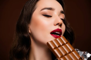 Kahverengi arka planda izole edilmiş, çarpıcı, tutkulu, çikolata ısıran bir kadının yakın plan fotoğrafı..