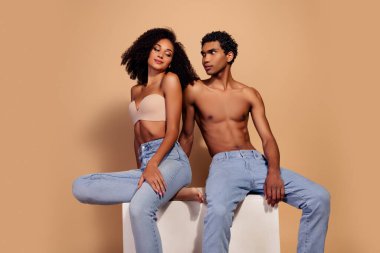 Yan yana oturan genç çift, bej bir arka planda romantizm ve zarafet sergiliyorlar. Sahne, stüdyo ortamında moda ve doğal güzelliği ön plana çıkarıyor..