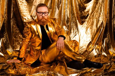 Retro altın takım elbiseli karizmatik bir adam, parlak altın arka planda, zarafet ve tarzın somutlaştırılmış, bayram veya parti temaları için mükemmel pozlar veriyor..