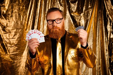 Altın takım elbiseli, sakallı, zarif, olgun bir adam poker kartları ve çip taşıyor, ışıldayan bir arka plana karşı şık ve karizmatik bir atmosfer yaratıyor..