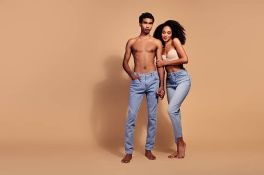 Genç bir çift güven içinde birbirlerine poz veriyor, birbirlerine sarılıyorlar. Bej rengi arka plan, şık görünüşlerini ve ilişkilerinin doğal güzelliklerini vurguluyor..